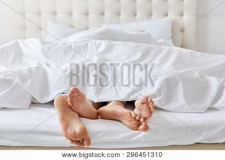 Horizontal Shot Of Feet Of Couple Under White Bedclothes In Bed At Bedroom, Enjoying Good Rest At Ho