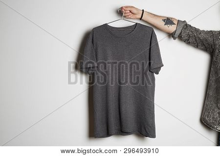 Tattooed Male Hand Holds Hang With Blank Gray T-shirt From Premium Thin Cotton On White Background. 