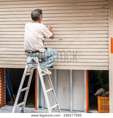 An Elderly Man Repairing Metal Blinds, Sitting On The Stairs . The View From The Back