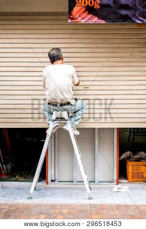 An Elderly Man Repairing Metal Blinds, Sitting On The Stairs . The View From The Back