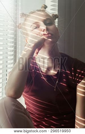 Serious Sad Young Woman With Hair Curlers On Head Sitting On Window Near Blinds