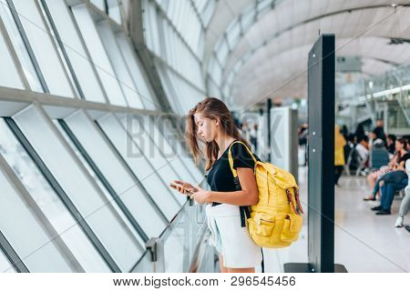 Teen girl using smarthphone while waiting for international flight in airport departure terminal. Young passenger with backpack travelling on airplane. Teenager tourizm abroad alone concept.