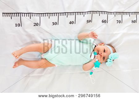A Three Month Baby In Mint Green Clothes Lying On A Bed On Which A Measuring Ruler For Growth Is Dra