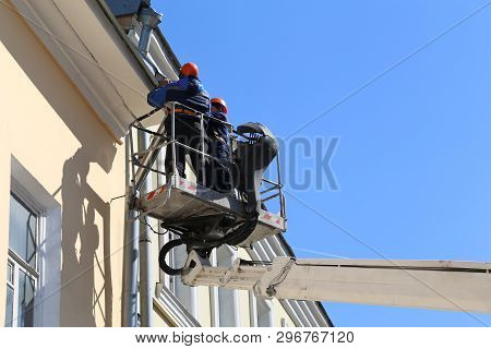 Lifting Platform With Workers Against The Blue Sky, Builders Are Repairing The Roof Gutter. Lifting 