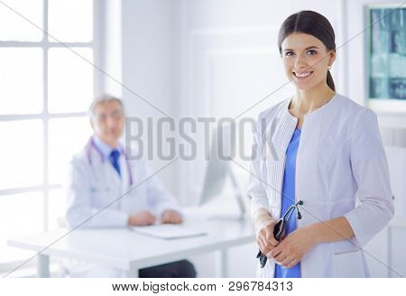 A doctor smiling at the camera with her male colleage in the back of the consulting room in hospital