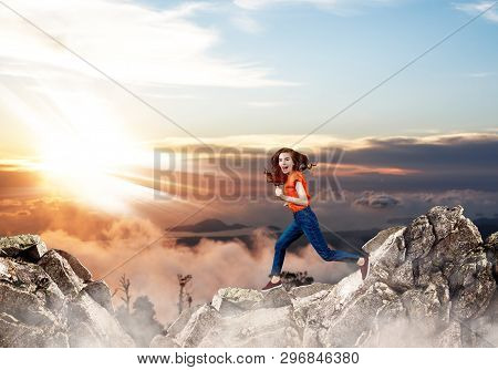 Redhead Woman Jumps Through The Gap Between Hills Over Cliff On Blue Sky Background.