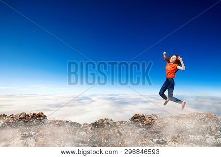 Redhead Woman Jumps Through The Gap Between Hills Over Cliff On Blue Sky Background.