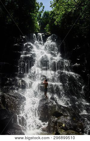 A Guy Is Standing Under A Waterfall. Beautiful Kanto Lampo Waterfall. Bali, Indonesia. Day Light