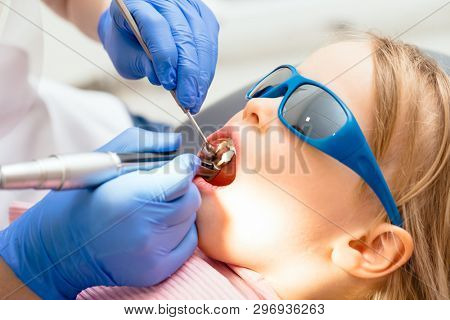 Dentist performing dental procedure to a little girl in pediatric dental clinic. Doctor using prophy brush on handpiece to polish filling. Calm child is sitting in a dental chair wearing sunglasses