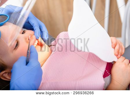 Dentist showing to little girl cured tooth in pediatric dental clinic. Child is sitting in a dental chair inspecting her teeth looking thru tooth-shaped mirror