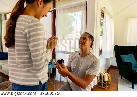 Young Man Proposing His Girlfriend With Engagement Ring At Home. Asian Woman Getting Surprised By He