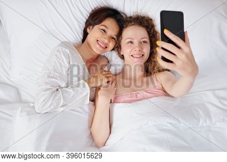Happy Pretty Young Multi-ethnic Lesbian Couple Lying In Bed And Taking Selfie