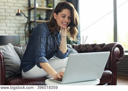 Smiling Young Woman Sitting On Sofa With Laptop Computer And Chating With Friends.