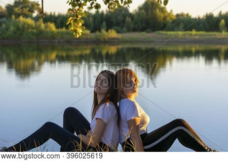 Two Young Women Are Resting In Summer By The River. Blonde And Brunette Are Back To Back In Nature. 