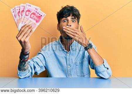 Young hispanic man holding yuan chinese banknotes sitting on the table covering mouth with hand, shocked and afraid for mistake. surprised expression 