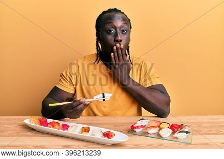 Handsome young black man eating sushi sitting on the table covering mouth with hand, shocked and afraid for mistake. surprised expression 