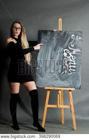 A Young Girl In A Black Dress And Hetaera, With Long Hair And Glasses, Points To A Chalkboard