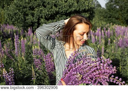 Smiling Beautiful Girl In Striped Blouse Posing With Lupine Armful And Her Hand Up On Backdrop Of Bl