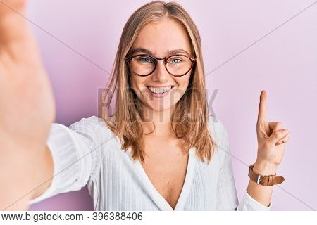 Beautiful blonde woman taking a selfie photo wearing glasses smiling with an idea or question pointing finger with happy face, number one 