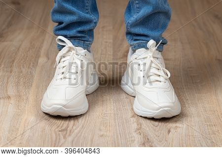 Standing Feet Shod In White Lace-up Chunky Sole Sneakers And Blue Jeans On The Brown Floor. Pair Of 