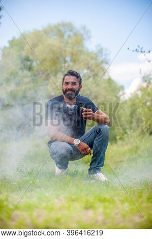 Stylish Bearded Man Exhaling Vapor From Vape In Nature. Electronic Cigarette.