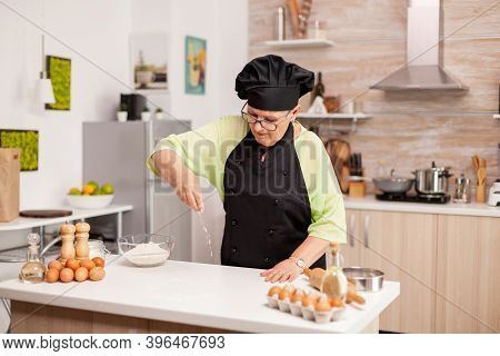 Woman Is Sifting Flour On Wooden Table In Modern Kitchen. Happy Elderly Chef With Uniform Sprinkling