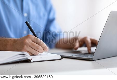 Unrecognizable Man Writing In Notepad And Using Laptop In Office, Taking Notes At Workplace, Using C