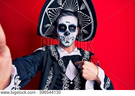 Young man wearing mexican day of the dead costume make selfie by the camera pointing finger to one self smiling happy and proud 