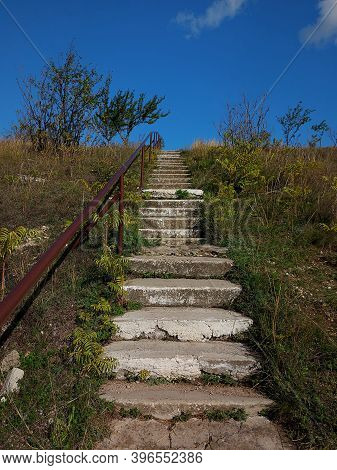 Old Concrete Stairs With Rusty Iron Pipe Handle Leading To The Blue Sky. High Hill With Trees And Gr