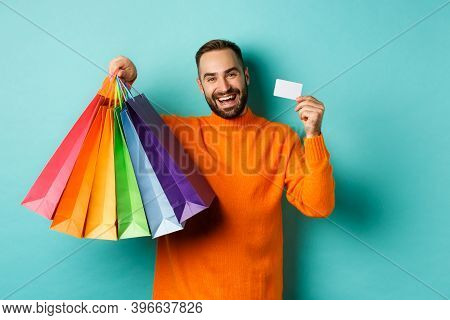 Happy Aduly Man Showing Credit Card And Shopping Bags, Standing Against Turquoise Background