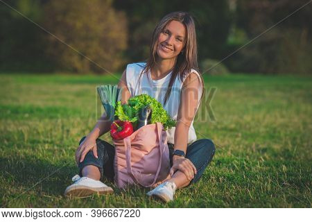 Beautiful Young Vegan Woman Sitting On The Grass With Pink Reusable Textile Shopping Bag With Fresh 