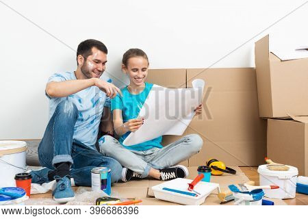 Happy Couple Sitting On Floor With Construction Blueprint. Home Remodeling And House Interior Redesi