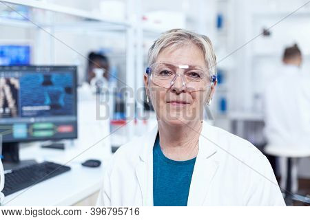 Close Up Portrait Of Biologist Looking At Camera In Sterile Laboratory. Elderly Scientist Wearing La