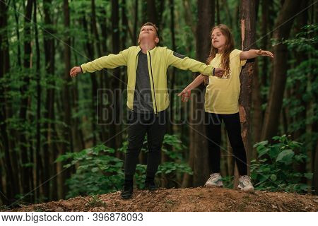 Little Boy And Girl With Raised Arms Enjoying The Fresh Air In Green Forest.
