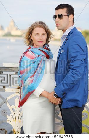 Stylish man in blue jacket and pregnant woman stand on bridge and hold hands.