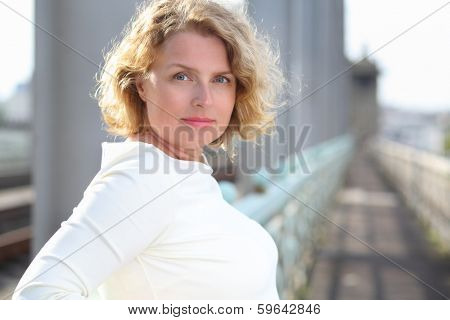 Happy beautiful blonde woman in white dress stands on bridge.
