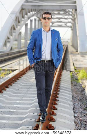 Handsome man in blue jacket and sunglasses stands on railroad near bridge.