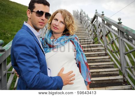 Pregnant woman in white stands on stairs and stylish man touches her belly outdoor. Focus on woman.