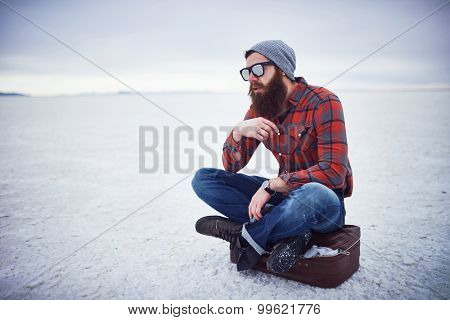 pensive contemplative hipster stroking awesome manly beard sitting on retro suitcase in salt flats all alone with copy space composition