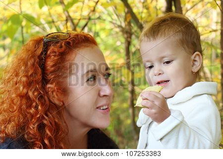Young Mother Talks With Her Little Daughter With Yellow Leaf In Hand At Autumn. Focus On Girl