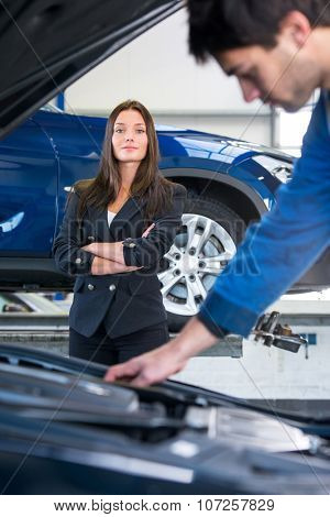 A business woman waits confidently, whilst a service mechanic is looking at the engine under the hood, examining her car in a garage