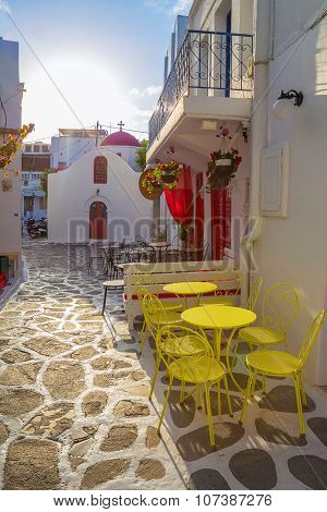 Mykonos streetview at sunrise with chapel and yellow chairs and tables, Greece