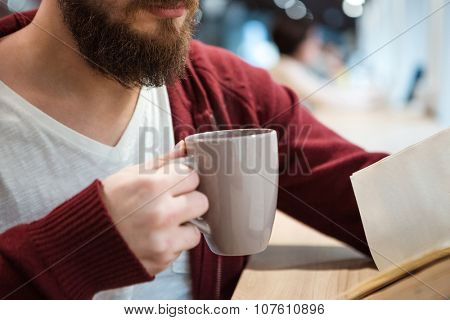 Young man with beard holding a cup of hot drink and reading