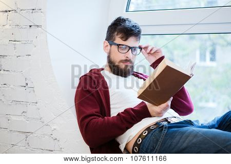 Young handsome man in glasses and brown hoodie reading on the window sill