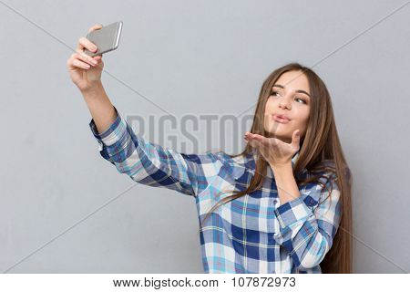 Beautiful happy girl with long hair in checkered shirt making selfie using cellphone sending an air kiss 