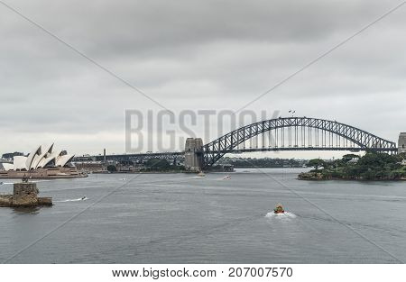 Sydney Australia - March 21 2017: Sydney Harbor bridge Opera House and Denison Fort under gray rainy sky. Darker gray water in front with small boats drawing white lines.