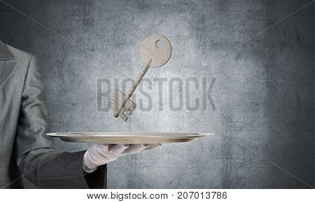 Closeup of waiter's hand in white glove presenting stone key symbol on metal tray with gray wall on background. 3D rendering.