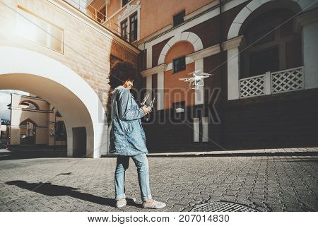 Cheerful young black lady in jean jacket is driving flying drone while standing on pavement stones near architectural arch and holding remote controller for operating quadrupter sunny autumn day
