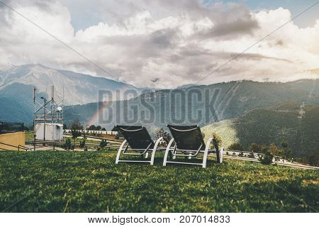 Two empty daybeds on grass glade and stunning rainbow mountains landscape behind; void recliners on autumn meadow with dramatic iris leaping over the hills in the background sunny day with rain
