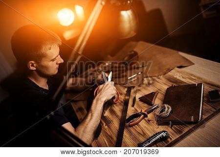 Working process of the leather belt in the leather workshop. Man holding crafting tool and working. Tanner in old tannery. Wooden table background. Warm Light for text and design.